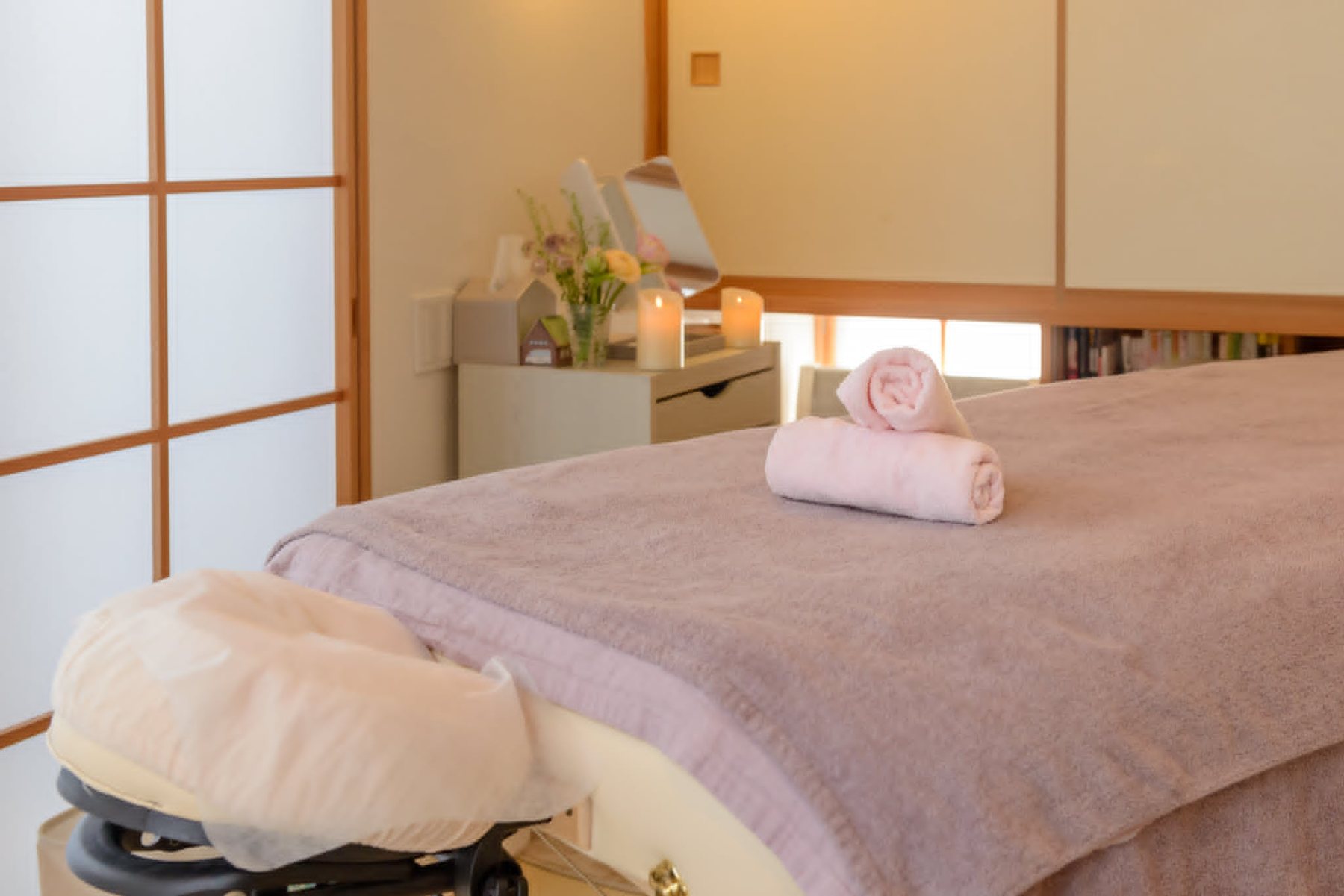 障子から光の入る和室。施術ベッドの上にたたまれた桜色のタオル、奥に花とキャンドル