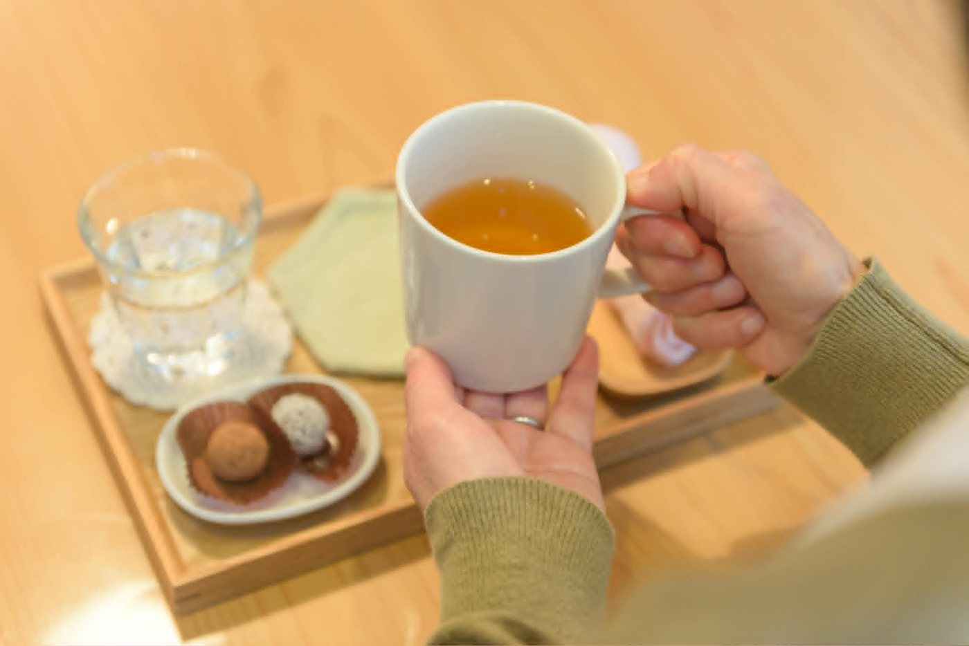 木のトレーにのった温かいお茶と小さなお菓子を、両手で包むように持っているところ